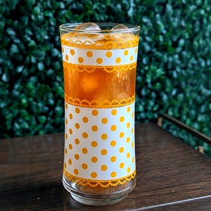 Puede incluir: Un vaso alto y transparente lleno de un líquido color ámbar y cubitos de hielo. El vaso presenta una banda blanca decorada con lunares amarillos y un patrón de encaje festoneado amarillo en los bordes superior e inferior.