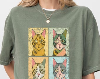 May include: An olive green short-sleeve t-shirt featuring a central graphic print of four cat portraits arranged in a square grid. Each cat is depicted in a different color palette, including yellow, pink, green, and blue, with a distressed texture.