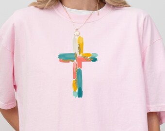 May include: A light pink short-sleeved t-shirt featuring a central graphic of a cross composed of multi-colored paint brush strokes in shades of teal, orange, yellow, and pink. The shirt has a crew neckline and a relaxed, loose-fitting silhouette.
