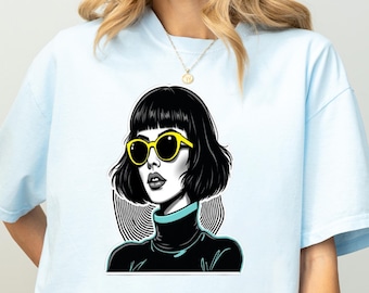 May include: A light blue t-shirt featuring a graphic illustration of a person with a black bob haircut, wearing bright yellow sunglasses and a black turtleneck sweater with a teal collar, set against a circular background of black lines.