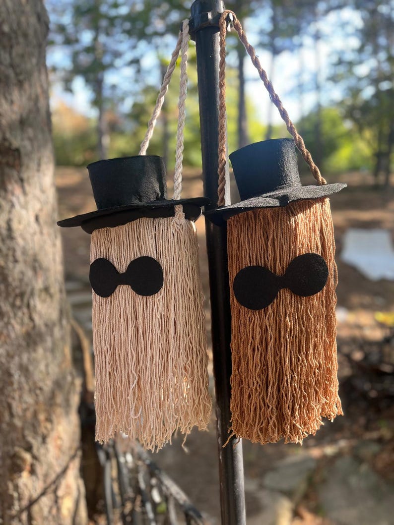 May include: Two hanging Halloween decorations. Each has a black top hat, a fringe body, and black eye accents. One is beige, the other is brown. They hang from a black metal pole with rope.