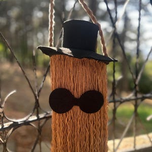May include: A handmade, whimsical shoulder bag with a black top hat and a long, fringed body in shades of brown. The bag features a pair of black, circular eye-like shapes. The bag is suspended by a braided rope strap.