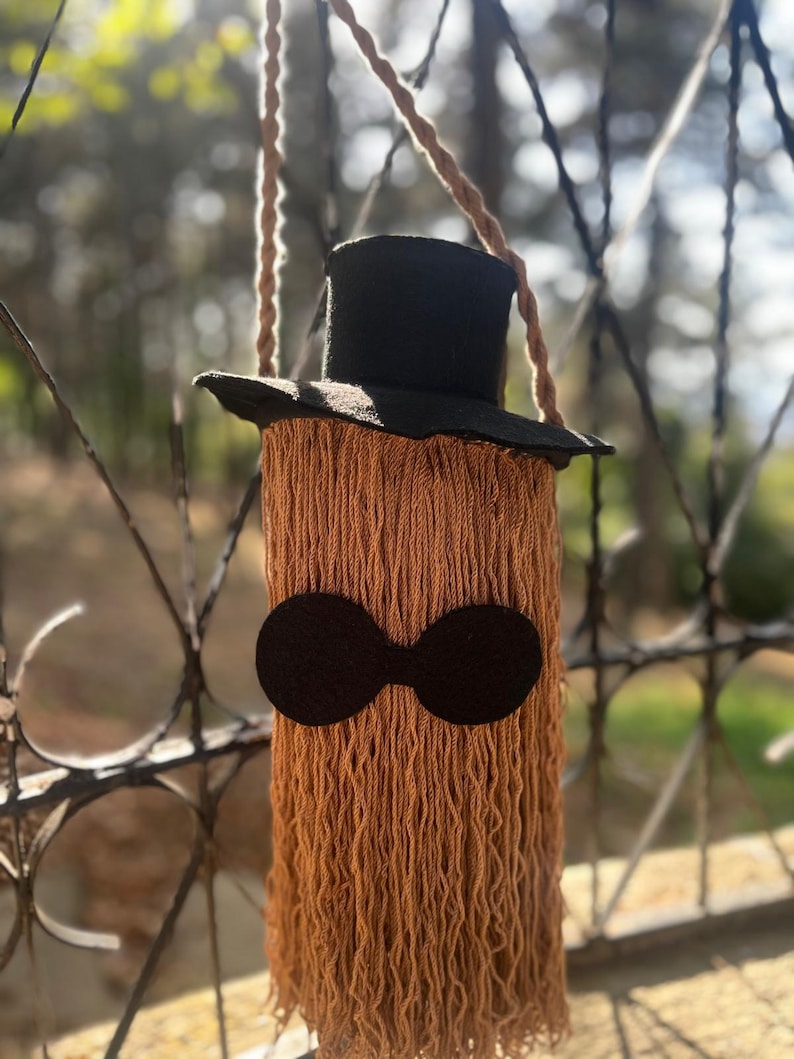 May include: A handmade, whimsical shoulder bag with a black top hat and a long, fringed body in shades of brown. The bag features a pair of black, circular eye-like shapes. The bag is suspended by a braided rope strap.