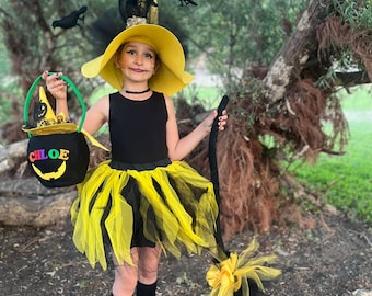 May include: A child dressed in a yellow and black witch costume, holding a broom and a trick-or-treat bag. The costume includes a wide-brimmed yellow hat, a black top, and a layered tulle skirt. The bag has the name "CHLOE" on it.