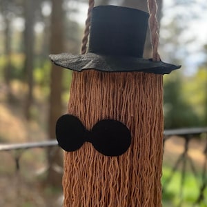 May include: A whimsical, handmade wall hanging in the shape of a character. It features a black top hat, a pair of black oval-shaped glasses, and a body made of brown yarn. The text "Style B" is in a black rectangle in the upper left corner.