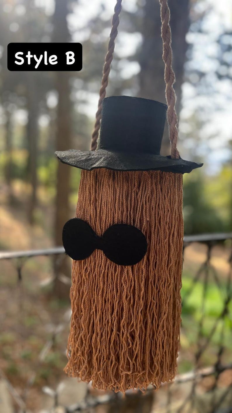 May include: A whimsical, handmade wall hanging in the shape of a character. It features a black top hat, a pair of black oval-shaped glasses, and a body made of brown yarn. The text "Style B" is in a black rectangle in the upper left corner.
