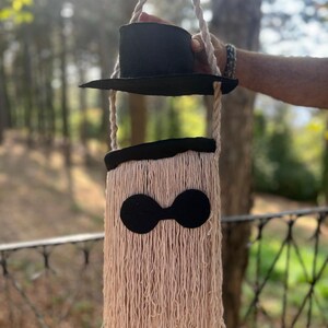 May include: A whimsical, handmade shoulder bag with a black top hat design. The bag features a beige, fringed body with a black, rounded shape detail. The bag is suspended by a rope handle.