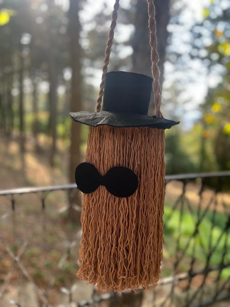 May include: A whimsical, handmade bag resembling a character with a black top hat and sunglasses. The bag is made of brown yarn, with a long, fringed body. The hat and sunglasses are black. The bag hangs from a twisted rope.
