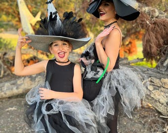 May include: Two people dressed in witch costumes. The costumes include black dresses, tulle skirts, and large hats adorned with feathers and bats. The hats are gray and black. The background is a natural outdoor setting with trees.