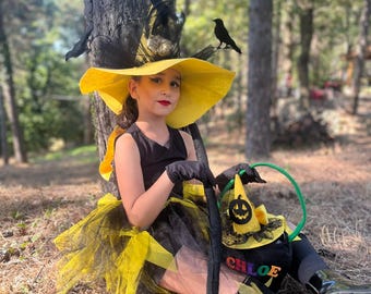 May include: A young person dressed in a witch costume, wearing a large yellow hat with black accents and a black dress. The costume includes a broom and a trick-or-treat bag with the name "Chloe" on it. The background is a forest.