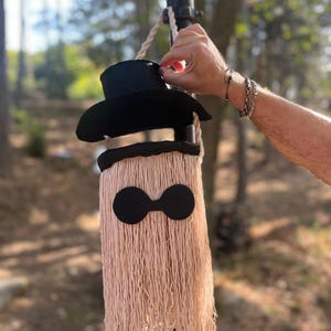 May include: A whimsical, handmade wall hanging featuring a black top hat and a fringed body in a light peach color. The design includes two large, black circular shapes, resembling eyes. The piece is suspended from a rope, creating a unique decorative item.
