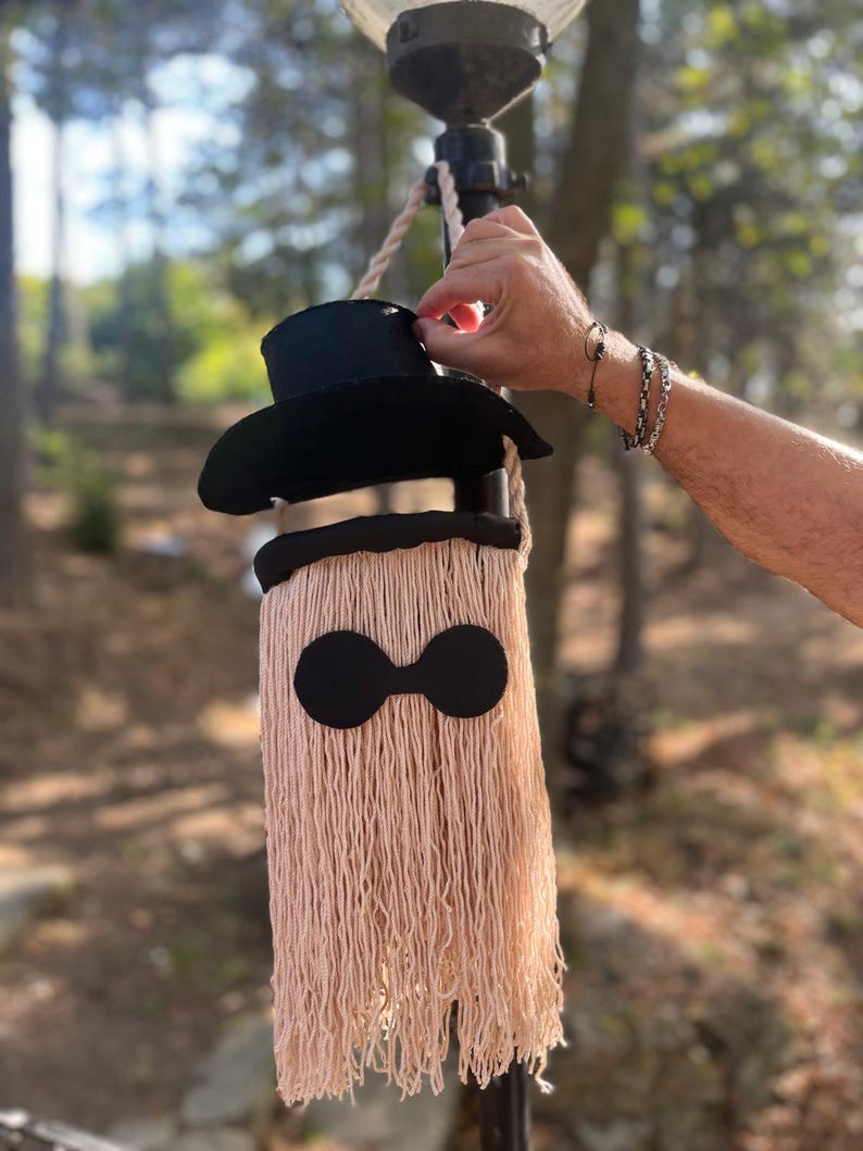 May include: A whimsical, handmade wall hanging featuring a black top hat and a fringed body in a light peach color. The design includes two large, black circular shapes, resembling eyes. The piece is suspended from a rope, creating a unique decorative item.