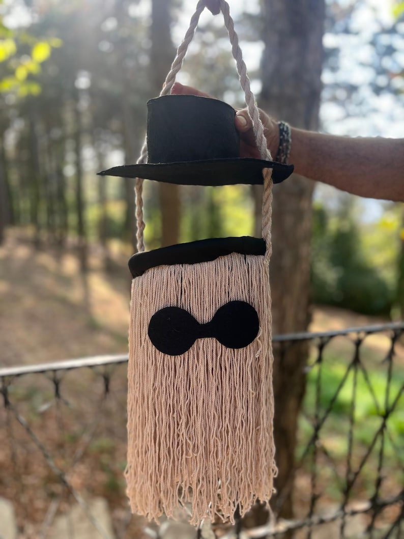May include: A whimsical, handmade shoulder bag with a black top hat design. The bag features a beige, fringed body with a black, rounded detail and a pair of black circles. The bag is suspended by a rope handle.