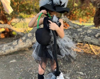 May include: A child's witch costume featuring a black dress, a large black hat with white tulle, and a matching broom. The costume includes a black bag with a green handle and black boots. The outfit is designed for Halloween or costume parties.