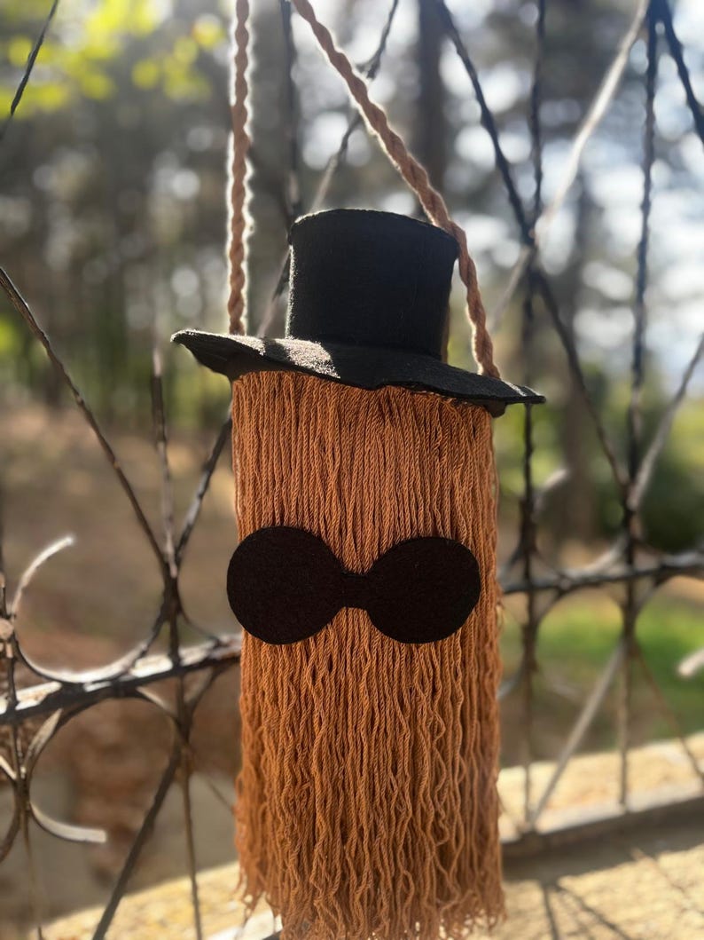 May include: A handmade, whimsical shoulder bag with a black top hat and a long, fringed body in shades of brown. The bag features a pair of black, circular eye-like shapes. The bag is suspended by a braided rope strap.