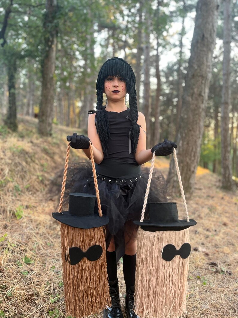 May include: A child in a black costume with a black wig and braids, holding two decorative broom-shaped bags. The bags are made of brown and tan yarn, with black hat tops and bow tie accents. The background is a forest.