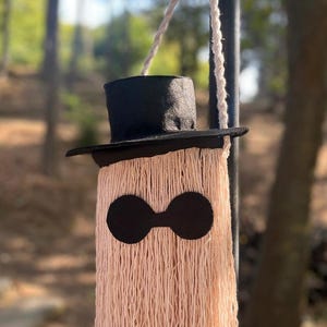 May include: A whimsical, handmade wall hanging in the shape of a ghost. It features a black top hat, black eye-shaped cutouts, and a body made of long, peach-colored yarn. The text "Style A" is in a black box in the upper left corner.