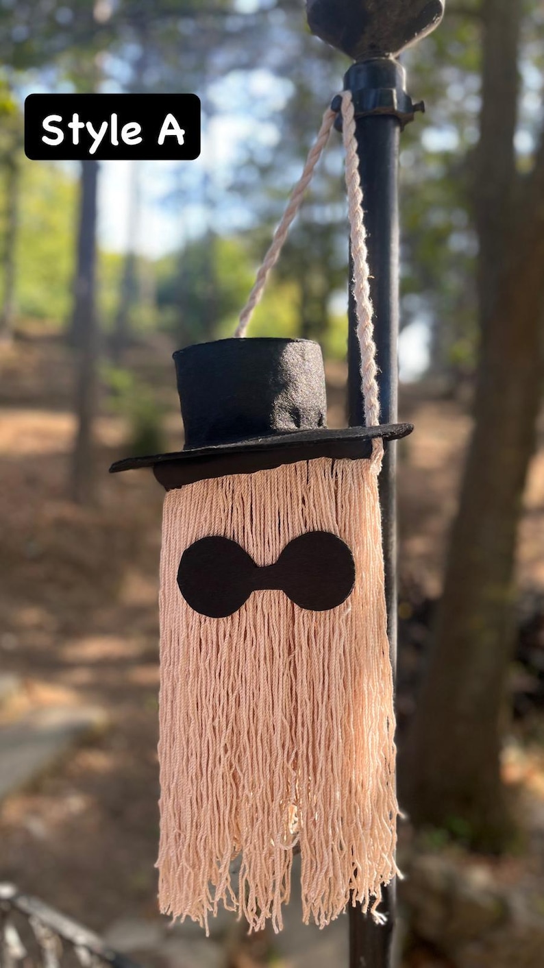 May include: A whimsical, handmade wall hanging in the shape of a ghost. It features a black top hat, black eye-shaped cutouts, and a body made of long, peach-colored yarn. The text "Style A" is in a black box in the upper left corner.