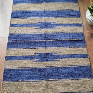 May include: A blue and beige striped rug with a geometric pattern. The rug has fringe on all sides.