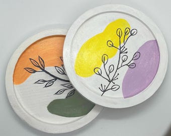 May include: Two white, round coasters with painted abstract designs. One coaster features orange and green shapes with a black leaf design. The other has yellow and purple shapes with a black leaf design. These coasters are perfect for adding a touch of art to your home decor.