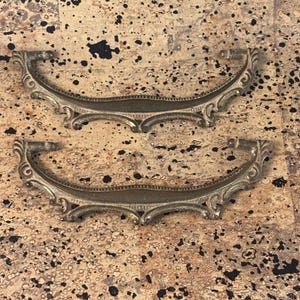 May include: Two antique brass drawer pulls with ornate, curved designs. The pulls have a decorative, scroll-like pattern and are set against a speckled, neutral-toned background. These vintage hardware pieces are ideal for furniture restoration projects.