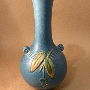 May include: A blue ceramic vase with a bulbous base and a slender neck. The vase features two small handles and a decorative relief of leaves and berries in shades of green, orange, and blue. The vase is approximately 8 inches tall.