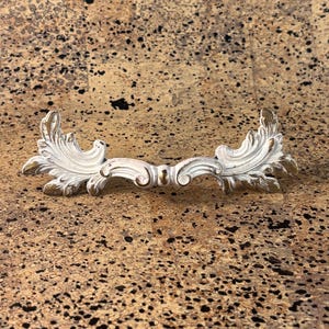 May include: A white and gold decorative furniture handle with an ornate, symmetrical design. The handle features a curved shape with detailed leaf and scroll accents. The hardware is likely made of metal and is designed for a cabinet or drawer.