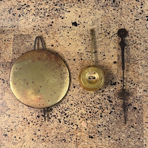 May include: Three antique clock parts: a large, round, brass pendulum, a brass bob, and a black metal clock hand. The pendulum has a curved top and a long, thin rod. The bob is round with a keyhole. The clock hand is ornate.