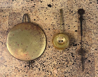 May include: Three antique clock parts: a large, round, brass pendulum, a brass bob, and a black metal clock hand. The pendulum has a curved top and a long, thin rod. The bob is round with a keyhole. The clock hand is ornate.