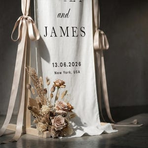 May include: A wedding welcome sign with the names "and JAMES" printed in black on a white fabric. The date "13.06.2026" and location "New York, USA" are also printed. The sign is decorated with dried flowers and beige ribbons.