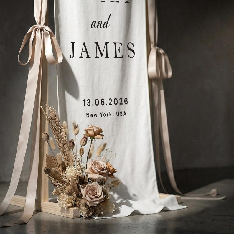 May include: A wedding welcome sign with the names "and JAMES" printed in black on a white fabric. The date "13.06.2026" and location "New York, USA" are also printed. The sign is decorated with dried flowers and beige ribbons.