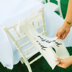 May include: Cream-colored chair with a white "Reserved" banner. The banner is tied with a bow and has a frayed edge. The text "Reserved" is written in black script. The chair is on a green surface.