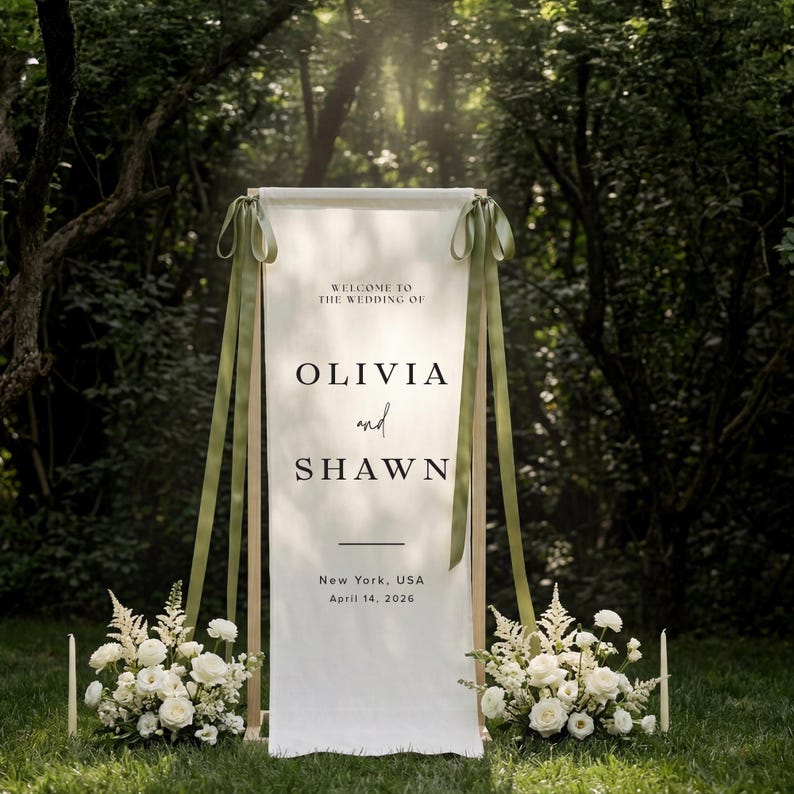 May include: A wedding welcome sign with the names Olivia and Shawn, set in a natural outdoor setting. The sign is white with green ribbons and wooden supports. The text includes the wedding date and location, surrounded by floral arrangements and candles.