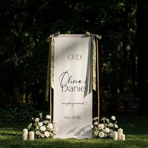 May include: A white wedding welcome sign with the names "Olivia & Daniel" in elegant script, framed by a copper-colored structure. The sign includes the text "are getting married" and the date "May 2026". Green ribbons and floral arrangements add decorative touches.