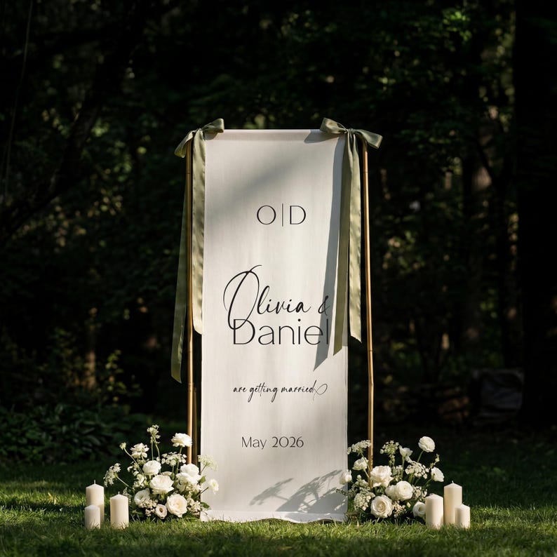 May include: A white wedding welcome sign with the names "Olivia & Daniel" in elegant script, framed by a copper-colored structure. The sign includes the text "are getting married" and the date "May 2026". Green ribbons and floral arrangements add decorative touches.
