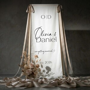 May include: A cream-colored wedding banner with the names "Olivia & Daniel" in elegant script. The banner is adorned with beige ribbons and a floral arrangement at the base. The text "are getting married" and "May 2026" are also displayed.