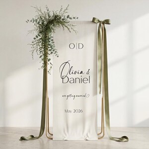 K&ouml;nnte beinhalten: Eine wei&szlig;e Hochzeitsanzeige mit den Namen "Olivia & Daniel" in Schreibschrift, dem Text "are getting married" und dem Datum "Mai 2026". Mit goldenem Rahmen, olivgr&uuml;nen B&auml;ndern und gr&uuml;nen Dekorationen.