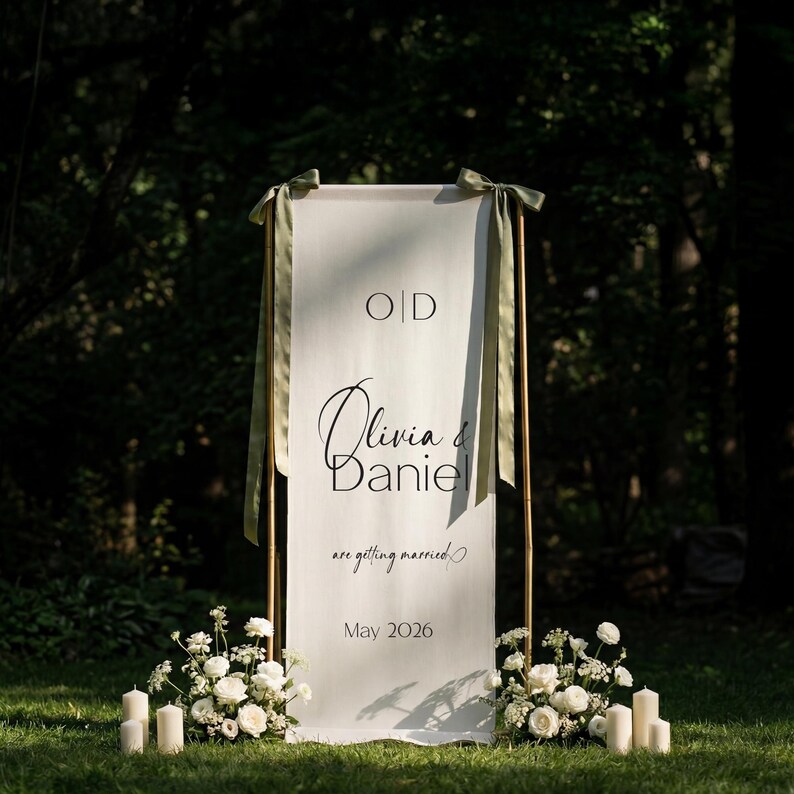May include: A white wedding welcome sign with the names "Olivia & Daniel" in elegant script. The sign is framed by a copper-colored structure and adorned with green ribbons. The date "May 2026" is also displayed. White flowers and candles are at the base.
