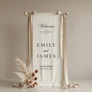 May include: A wedding welcome sign with the names Emily and James, and the date 06/13/2026. The sign is cream-colored with gold accents and ribbons. Dried flowers and pampas grass are at the base of the sign.