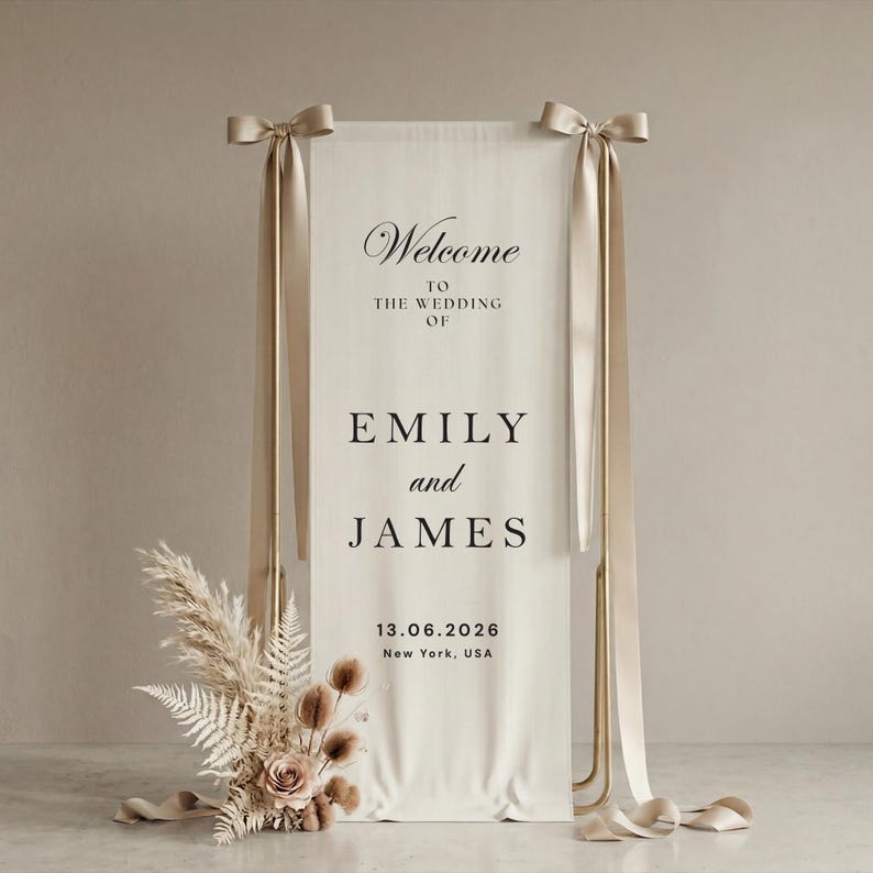 May include: A wedding welcome sign with the names Emily and James, and the date 06/13/2026. The sign is cream-colored with gold accents and ribbons. Dried flowers and pampas grass are at the base of the sign.