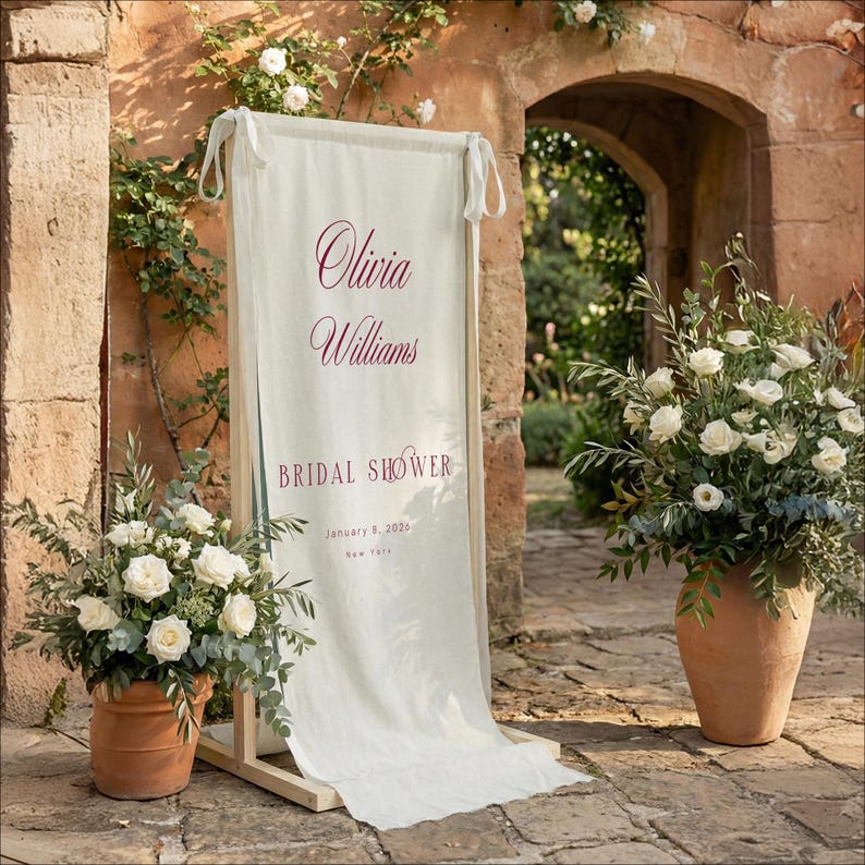 以下が含まれることがあります： ブルゴーニュ色の筆記体で「Olivia Williams BRIDAL SHOWER」と書かれたクリーム色の看板。看板は木製のフレームにあり、両側にテラコッタの鉢に入った白い花があります。日付は2026年1月9日、ニューヨークです。