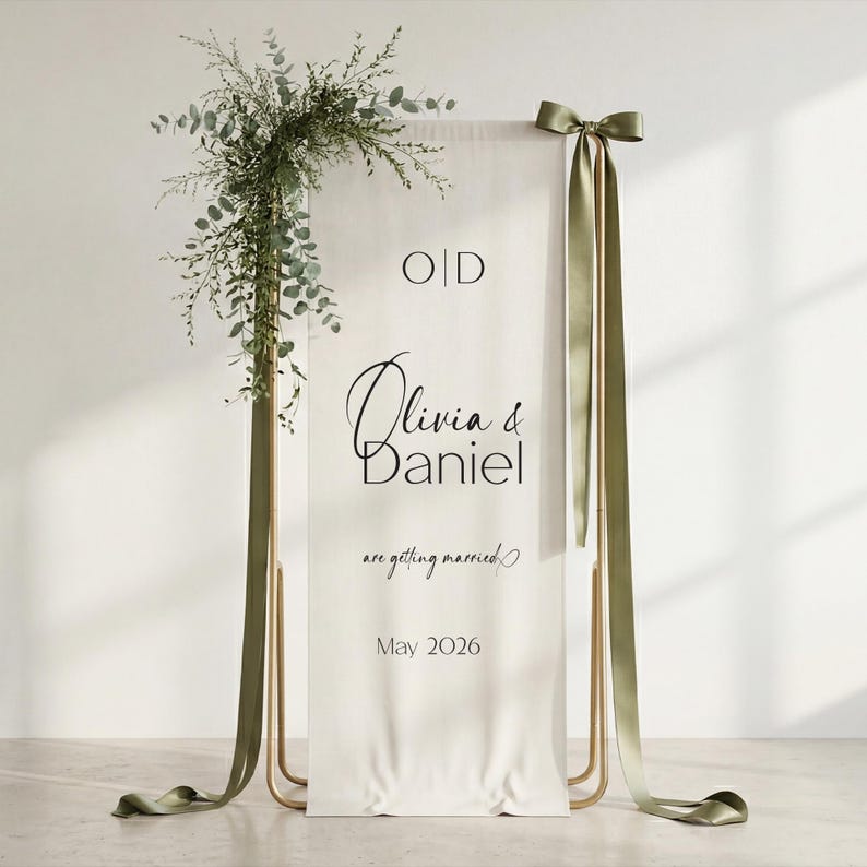 Personalized linen wedding welcome sign with olive green satin ribbons displayed on a gold stand, minimalist fabric wedding sign styled for garden ceremony entrance, boho wedding decor with natural light and greenery, custom wedding welcome banner with serif typography, elegant neutral linen wedding sign for outdoor wedding, bridal shower backdrop, modern wedding signage with soft draped fabric and organic styling