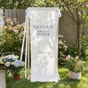 以下が含まれることがあります： ネイビーブルーの「OLIVIA'S BRIDAL SHOWER」の文字が入った白い布バナー。バナーには日付「2026年10月3日」と「NEW YORK」も含まれています。バナーは白いリボンが付いた木製のフレームから吊り下げられています。