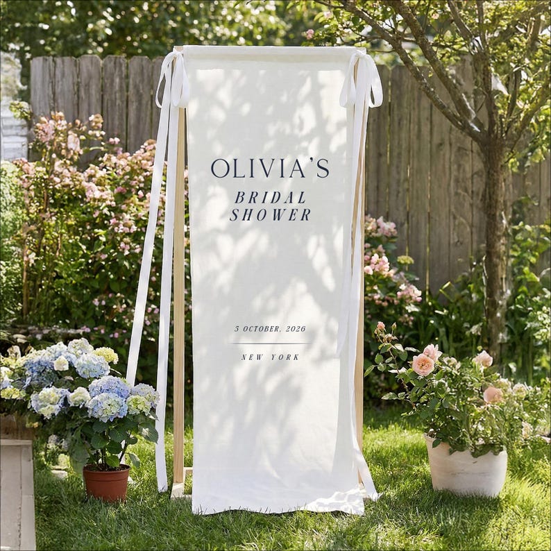 以下が含まれることがあります： ネイビーブルーの「OLIVIA'S BRIDAL SHOWER」の文字が入った白い布バナー。バナーには日付「2026年10月3日」と「NEW YORK」も含まれています。バナーは白いリボンが付いた木製のフレームから吊り下げられています。