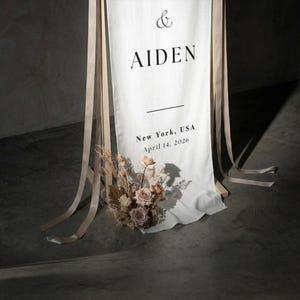 May include: A white fabric wedding banner with the names "& AIDEN" in black. The banner includes the text "New York, USA April 14, 2026". The banner is decorated with a floral arrangement and hanging ribbons.
