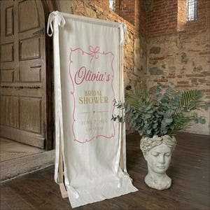 以下が含まれることがあります： 「Olivia's Bridal Shower」とピンク色の文字で書かれたクリーム色のバナー。日付は2026年6月7日、場所はラスベガス。バナーは木製のフレームと白いリボンで支えられています。頭の形をした装飾的なプランターには緑が植えられています。