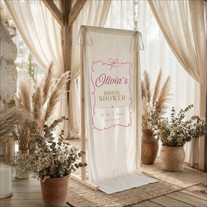 以下が含まれることがあります： 「Olivia's Bridal Shower」と書かれたピンク色の文字と、2026年6月7日の日付がラスベガスで表示された、クリーム色のブライダルシャワーの看板。看板は木製のフレームにあり、乾燥した植物や花に囲まれています。