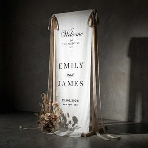 May include: A wedding welcome sign with the names "Emily and James" printed in black on a white fabric banner. The banner is suspended from a wooden frame and adorned with beige ribbons. A floral arrangement sits at the base.