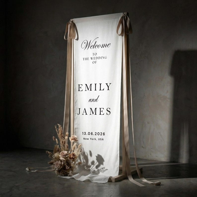 May include: A wedding welcome sign with the names "Emily and James" printed in black on a white fabric banner. The banner is suspended from a wooden frame and adorned with beige ribbons. A floral arrangement sits at the base.