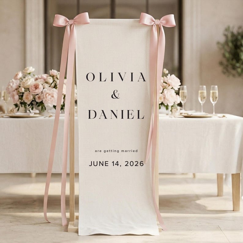 May include: A cream-colored wedding welcome sign with the names Olivia & Daniel and the date June 14, 2026. The sign is adorned with pink bows and ribbons. The sign is displayed at a wedding reception.